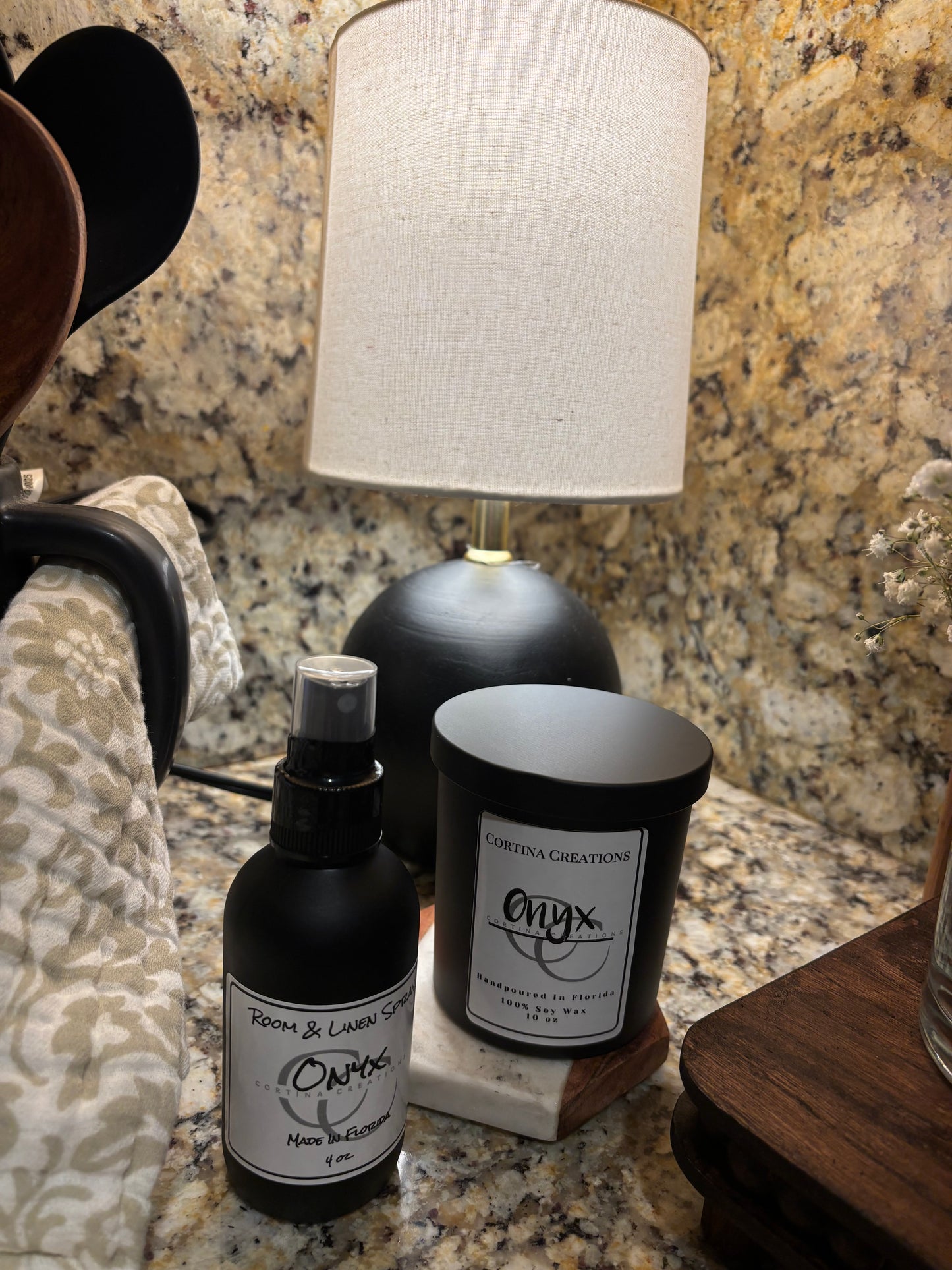 Two bottles one room spray one candle on a granite countertop with a lampshade in the foreground.